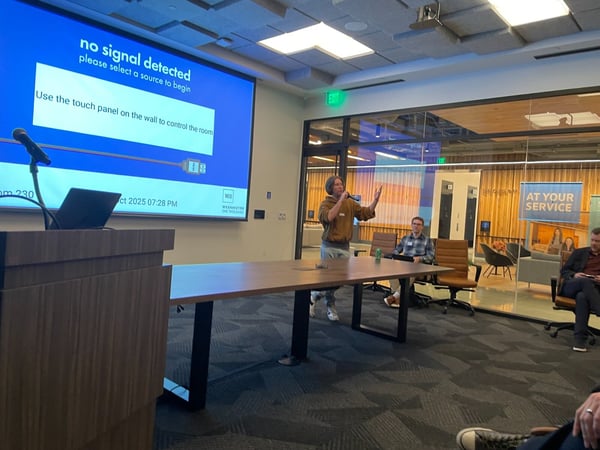 A candid photograph taken inside a modern conference room showing a man standing and speaking into a microphone during a presentation or meeting. A large screen behind him displays a blue 'no signal detected' message.