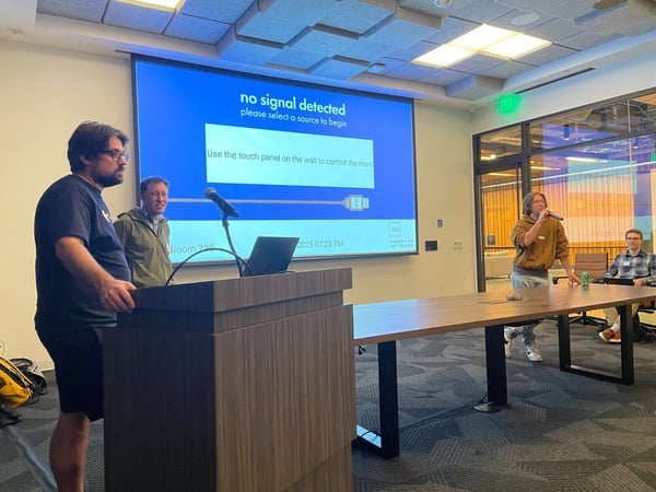 A photograph captures an indoor meeting or presentation taking place in a modern conference room, featuring four individuals gathered around a long table and a podium.