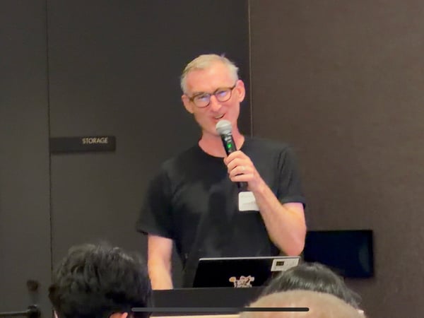 A man wearing glasses and a black t-shirt stands at a podium, holding a microphone and speaking to an audience.