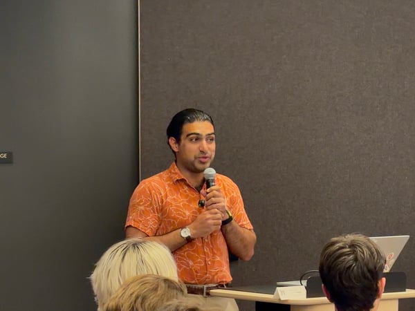 A man in an orange patterned shirt stands at a podium, speaking into a microphone during a presentation with audience members in the foreground.
