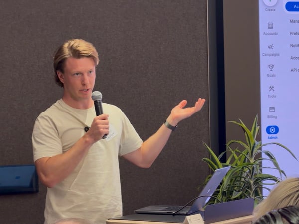 A young man is giving a presentation, holding a microphone and gesturing with his hand next to a large screen displaying a software menu.