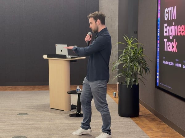 A man stands on a stage holding a microphone and gesturing during a presentation, with a screen behind him displaying 'GTM Engineering Track'.