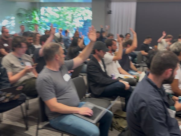 An indoor photograph of an audience seated in rows of chairs, with several people raising their hands during a presentation or Q&A session.