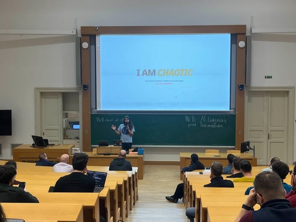 A photograph capturing a technical presentation taking place in a lecture hall, where a presenter stands beneath a large screen displaying the text "I AM CHAOTIC" to an audience seated in rows of wooden desks.