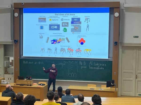 A presenter stands in a university lecture hall, gesturing toward a large projection screen that displays a technical slide about robotics and AI tools, while an audience is seated and observing.