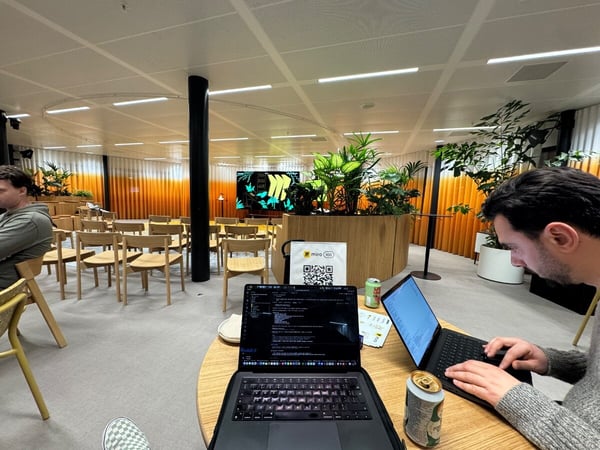 Two men are working intently on open laptops at a wooden table in a spacious, modern office auditorium featuring rows of empty chairs, large potted plants, and an orange-and-white gradient curtain backdrop.