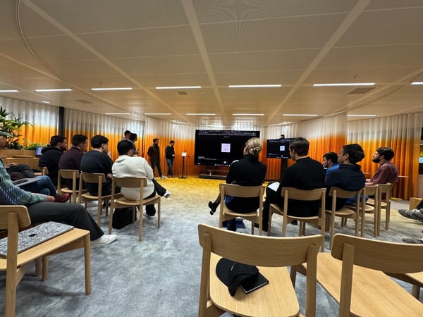 A candid photograph shows a group of people seated in a modern meeting room, facing two large screens where a presentation is being given by a few individuals standing near the displays.