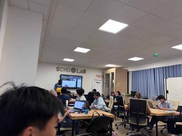 A candid photograph taken inside a modern classroom or workshop where several people are seated at wooden tables working on laptops, while a presenter stands near a large screen displaying content and the word 'SCHOOLAB' is visible on the wall.