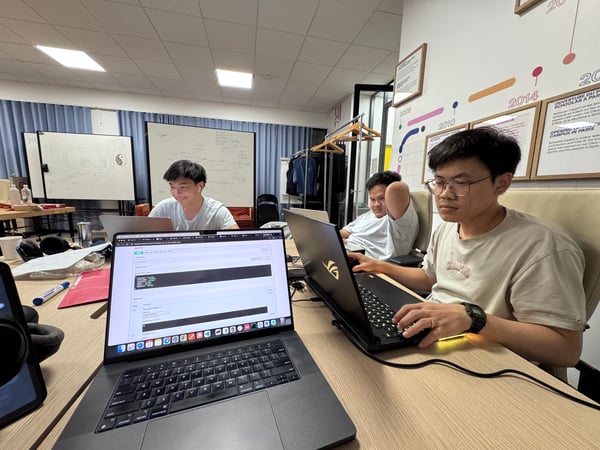 A candid photograph showing three young men working intently on their laptops at a long wooden desk in a collaborative indoor space, likely a classroom or co-working area.