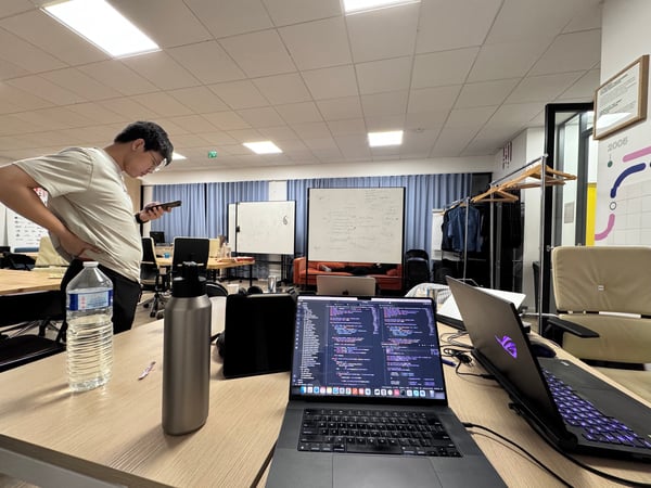 A candid indoor photograph of a modern office space, showing a desk with two open laptops displaying code in the foreground, and a man standing nearby checking his mobile phone.