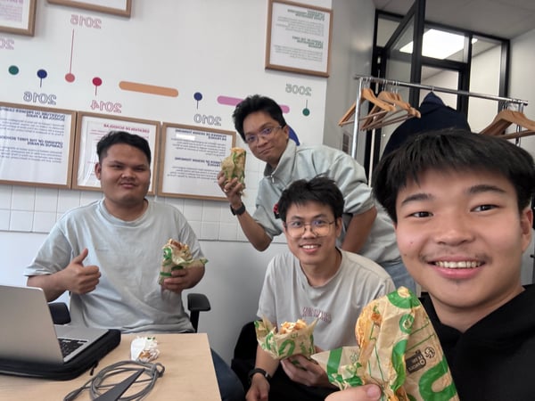 A close-up, casual photograph of four young men posing indoors, likely taking a selfie, while holding wrapped food items during a meal break.