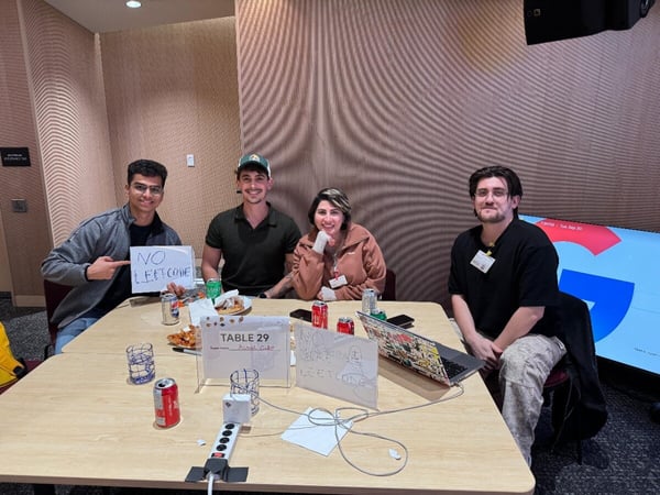 A photograph showing four young adults sitting around a table at an indoor event, likely a hackathon, with one person holding a sign that reads "NO LEETCODE."
