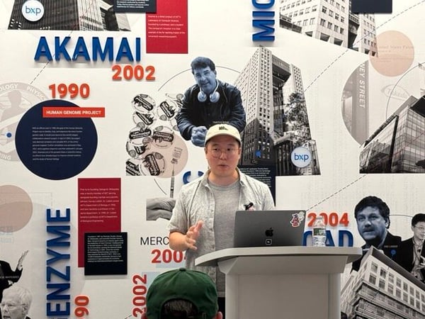 A man in a light-colored shirt and cap stands at a podium presenting with a laptop, set against a detailed graphic wall backdrop.