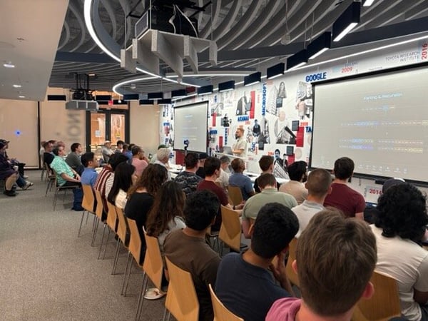 An indoor presentation setting showing an audience seated in rows of chairs watching a speaker standing near large projection screens.