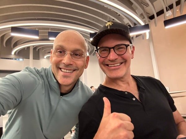 A selfie of two smiling men standing close together indoors under a modern, arched ceiling.