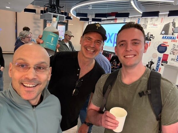 A casual indoor selfie of three smiling men, with one holding a bottle of Mamala rum and another holding a paper cup.