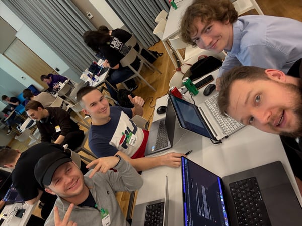 A candid group photo captures several young men smiling and posing for the camera while seated closely around white tables during an indoor coding event or hackathon. Many open laptops and beverage cans are visible on the cluttered tables, with other participants working in the background.