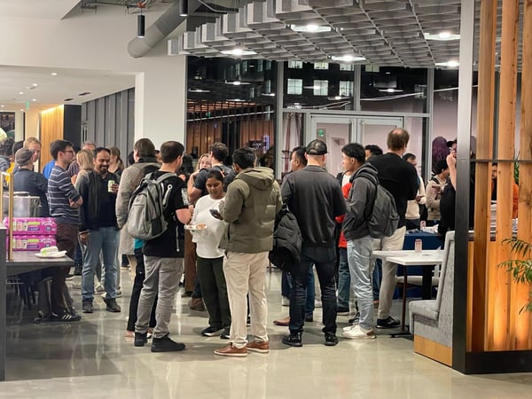 A candid photograph captures a large group of people mingling and socializing in a modern indoor space, likely an office or event venue, with some attendees holding food and drinks.