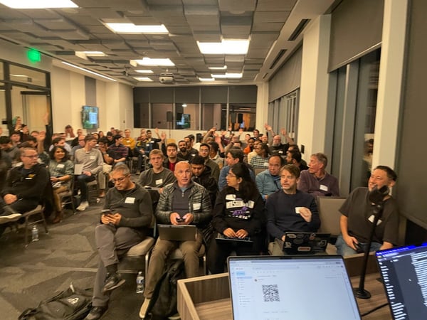 A candid photograph captures a large audience seated in a modern conference room during an event, with numerous attendees raising their hands, suggesting a Q&A session or vote.