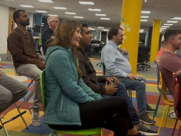 A candid indoor photo showing a diverse group of people seated in colorful chairs, attentively watching a presentation or event in a brightly decorated office space.