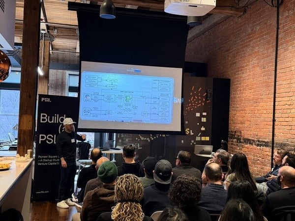 A man is giving a technical presentation to a seated audience in an industrial-style office space with an exposed brick wall and a projector screen.