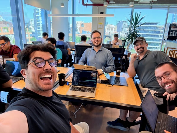 A group of men smiling for a selfie in a modern, sunlit office or co-working space with laptops open on a large wooden table.