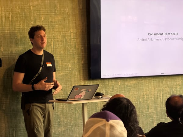 A speaker stands next to a projection screen displaying a slide about UI design while addressing an audience.