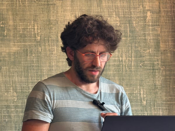 A man with curly hair and orange-rimmed glasses looking down at a laptop screen.