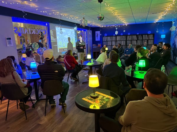 A man is giving a presentation to a seated audience in a dimly lit room decorated with string lights and colorful lamps. The presentation is projected onto a wall, showing a software interface or a game.