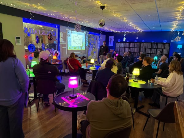 A group of people are gathered in a dimly lit, festively decorated lounge or bar, watching a presentation projected onto a wall.