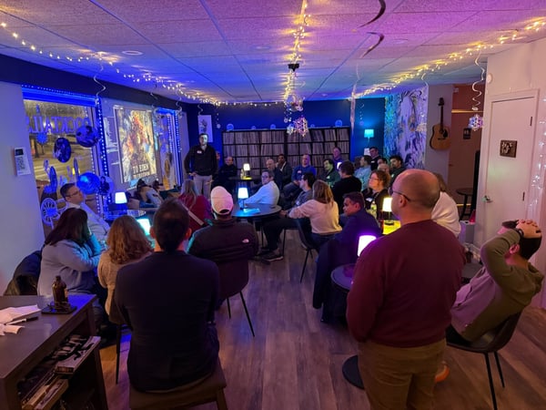A group of people are gathered in a dimly lit indoor space, likely a lounge or bar, watching a presentation or film projected onto a screen. The room is decorated with festive string lights and has a cozy, social atmosphere.