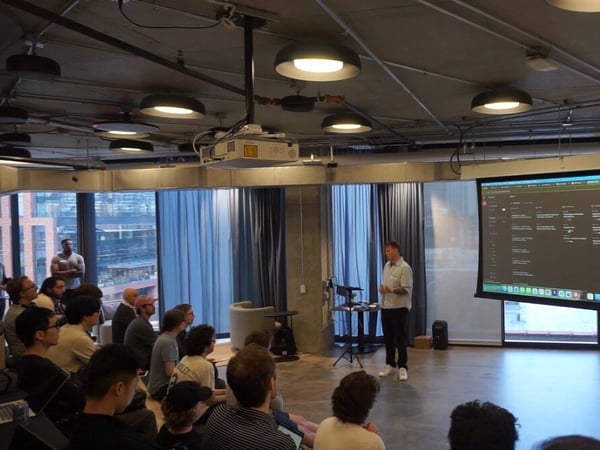 A man is giving a presentation to a group of people seated on the floor in a modern office or event space with a large projector screen.