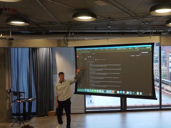 A man is giving a technical presentation in a modern office space, gesturing towards a large projection screen displaying a software interface.