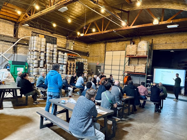A group of people sitting at picnic tables in a large brewery warehouse, watching a presentation given by a man standing next to a screen.