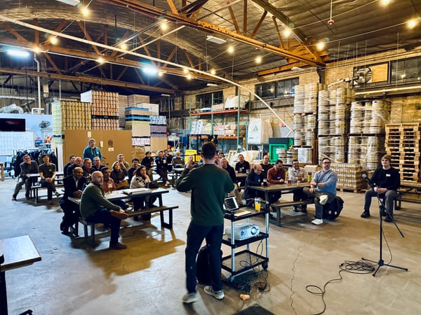 A man stands before an audience seated at picnic tables, giving a presentation in a large warehouse or brewery filled with stacks of kegs.