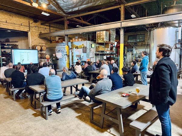 A group of people are gathered in a brewery or warehouse-style space for a presentation, with a speaker standing next to a large screen.