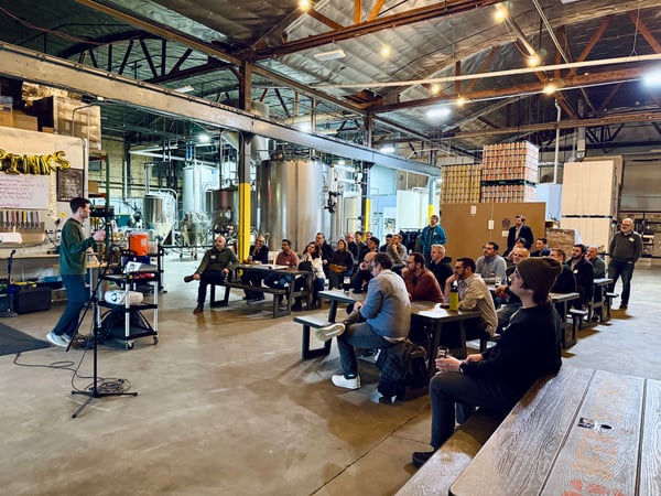 A man stands at a podium giving a presentation to a seated audience inside a large industrial brewery with stainless steel tanks and high ceilings.