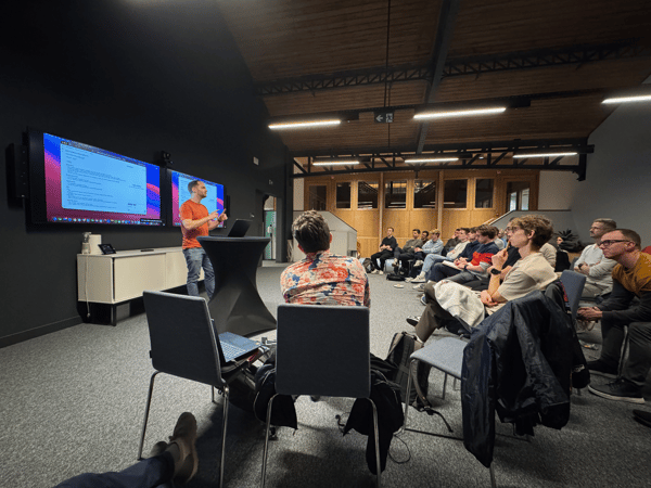 A candid photograph capturing an indoor technical presentation where a speaker in an orange shirt stands next to a laptop podium, addressing a large audience seated in rows facing two wall-mounted screens displaying code.