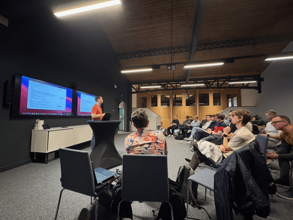 A man in an orange shirt is giving a presentation or lecture to a seated audience in a dimly lit, modern room featuring two large wall-mounted screens displaying text content. The audience, consisting mostly of young men, is seated in chairs and attentively watching the speaker.