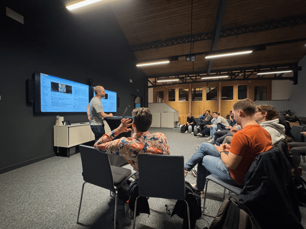 A candid photograph captures a technical presentation taking place in a dimly lit, modern conference room with dark walls and a high wooden ceiling, showing a presenter standing near dual screens while an audience is seated and observing.