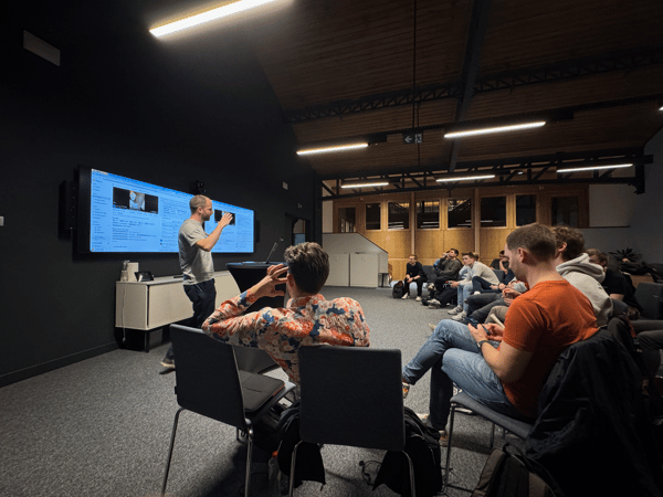 A photograph taken in a dimly lit, modern meeting space shows a man giving a presentation or demonstration in front of a large horizontal screen to a seated audience.