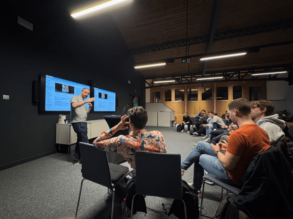 A man is giving a presentation or instruction in a dimly lit room, standing next to two large illuminated monitors displaying technical content, while a group of people sits and watches in the foreground and background.