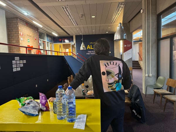 A person from behind stands in an indoor common area, wearing a black sweatshirt with a colorful abstract graphic. A bright yellow table in the foreground holds water bottles and snacks.