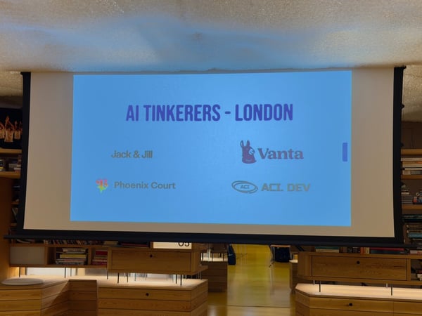 A photograph showing a large projection screen in an indoor event space, displaying the title "AI TINKERERS - LONDON" and several sponsor logos and names against a bright blue background. The screen is framed by wooden built-in shelving units filled with books on both sides.