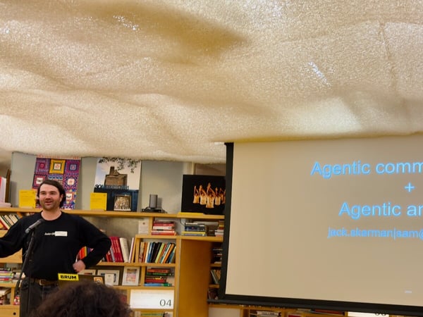 A photograph captures a man giving a presentation indoors, standing in front of a bookshelf while a projector screen displays the text 'Agentic communications + Agentic analytics'.