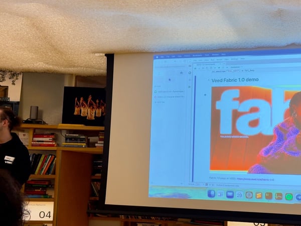 An indoor photograph capturing a presentation or demo where a man stands next to a large projection screen displaying content related to "Veed Fabric 1.0 demo," set against a backdrop of bookshelves and a textured ceiling.