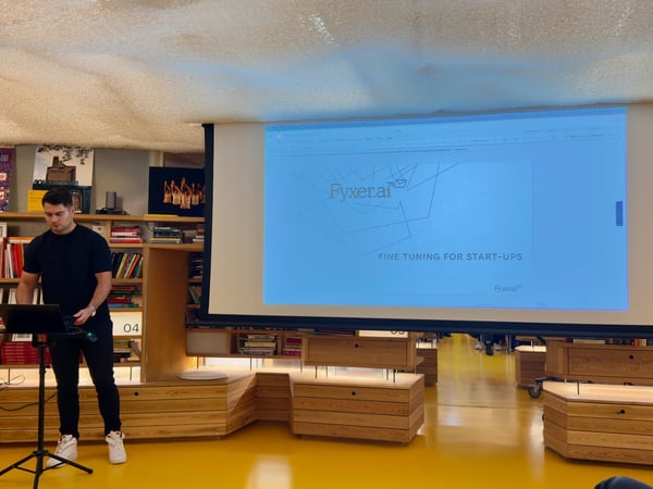 A man dressed in black is giving a presentation in a modern indoor venue characterized by a bright yellow floor and tiered wooden shelving units. The large projection screen behind him displays the title slide for "Fyxer.ai" concerning fine-tuning for start-ups.