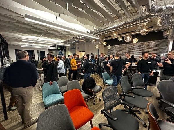 A photograph capturing a large group of people socializing and networking in a modern, brightly lit indoor event space or office, featuring distinctive wavy acoustic ceiling panels and various scattered lounge and office chairs in the foreground.