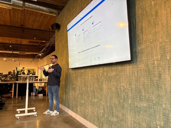 A man stands in a modern office space, giving a presentation next to a large television screen displaying a website for 'The InterviewDroid'.