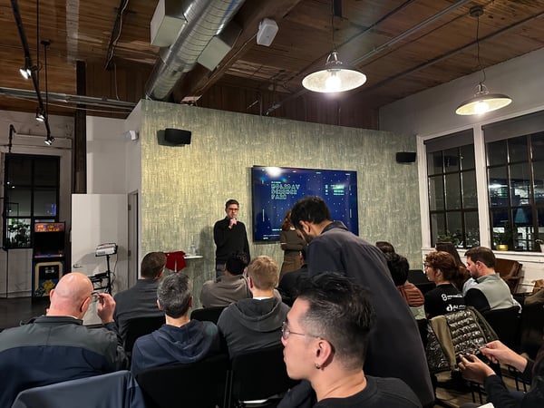 A photograph capturing an indoor presentation or meetup where a speaker, a man in a black sweater, stands near a large screen displaying text, addressing an audience seated in the foreground. The venue features an industrial aesthetic with exposed ductwork and wooden ceilings.